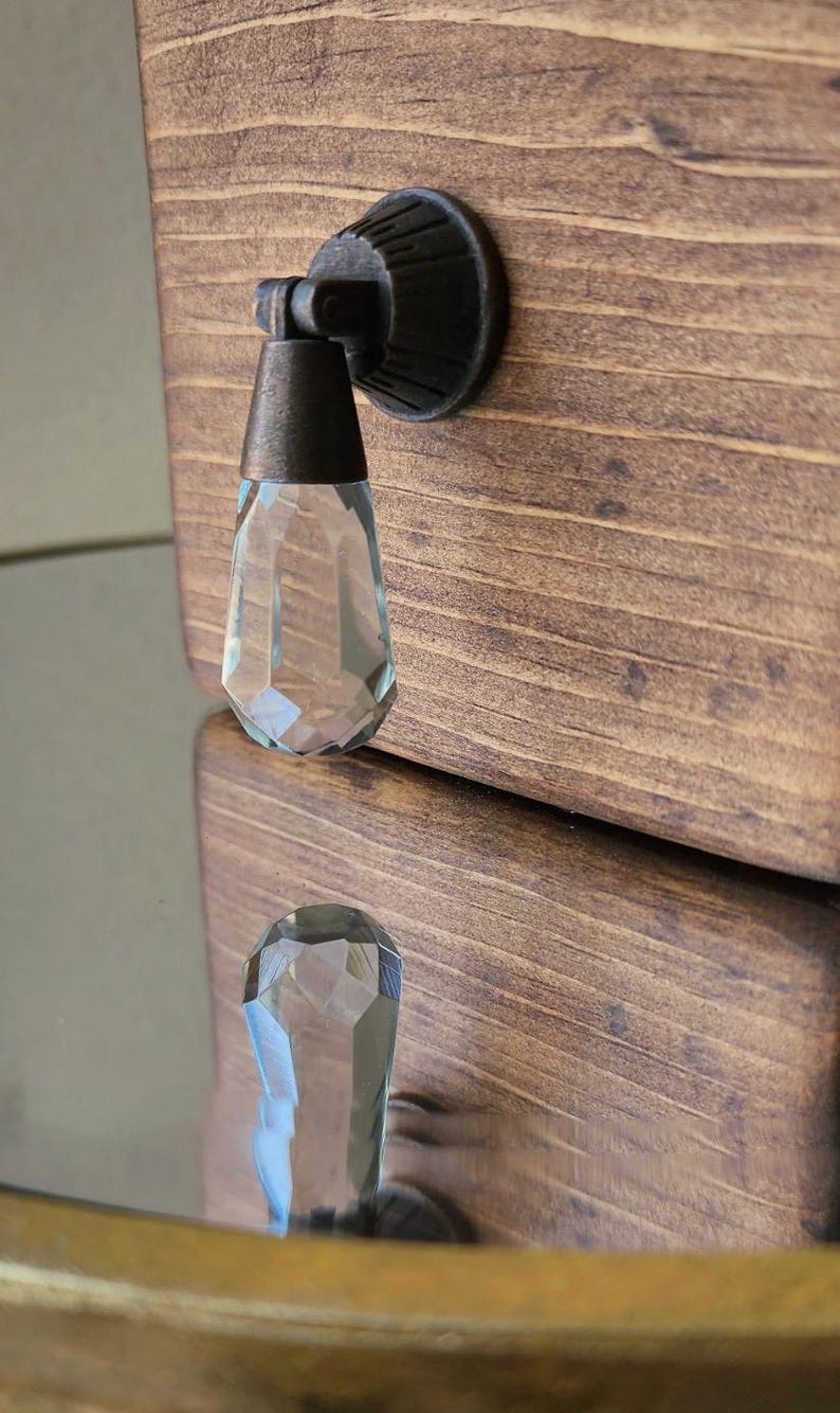 Peut inclure: Une poign&eacute;e de tiroir en verre transparent avec un design facett&eacute; et une base en m&eacute;tal bronze. La poign&eacute;e est mont&eacute;e sur un tiroir en bois.