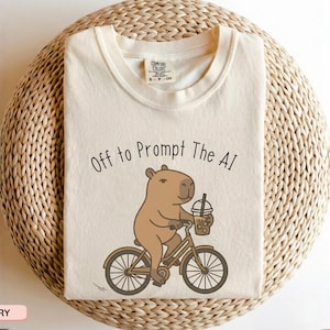 May include: Ivory-colored t-shirt featuring a cartoon capybara riding a bicycle and holding a bubble tea. The text "Off to Prompt The AI" is above the capybara. The shirt is displayed on a woven, circular surface.