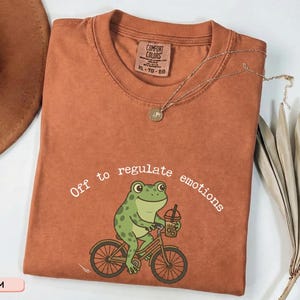Peut inclure: T-shirt de couleur terre cuite avec un col rond. Le t-shirt présente une grenouille de dessin animé à vélo et tenant une boisson, avec le texte "Off to regulate emotions". Un collier doré et une plante séchée sont également visibles.