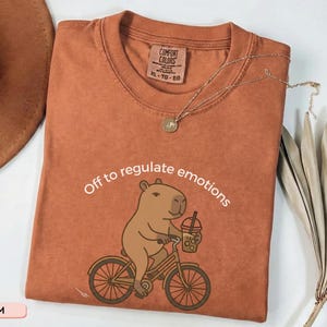 May include: Rust-coloured Comfort Colors t-shirt with the text "Off to regulate emotions" and a cartoon capybara riding a bicycle while holding a drink. A gold necklace and a hat are visible.