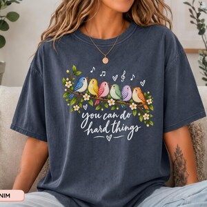 May include: A dark blue short-sleeve t-shirt featuring a graphic of six colorful birds perched on a flowering branch. Above the birds are musical notes and hearts. Below the branch, the text reads, you can do hard things, in white script.