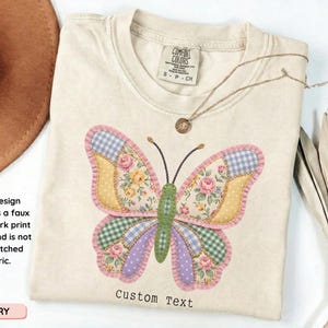 May include: Ivory-colored t-shirt with a large butterfly design. The butterfly features a patchwork print effect with floral, gingham, and polka dot patterns in pink, yellow, green, and blue. The text "Custom Text" is printed below the butterfly.