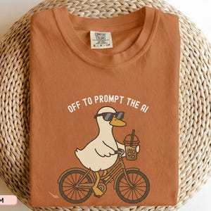 May include: A terracotta-colored t-shirt with a graphic of a duck wearing sunglasses and riding a bicycle while holding a drink. The text "OFF TO PROMPT THE AI" is above the duck. The shirt is displayed on a woven surface.