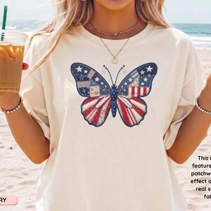 May include: Cream-coloured t-shirt with a butterfly design featuring a faux patchwork print. The butterfly's wings display a patriotic design in red, white, and blue. The shirt has a relaxed fit and short sleeves.