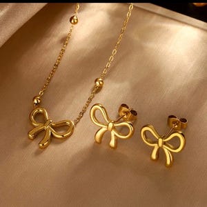 Peut inclure: Un ensemble de bijoux de couleur or comprenant un collier avec un pendentif en forme de nœud et des boucles d'oreilles assorties. Le collier a une chaîne délicate avec de petites perles rondes.