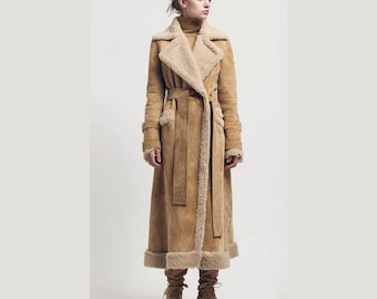 Könnte beinhalten: Ein langer, hellbrauner Wildledermantel mit Shearling-Futter und -Besatz. Der Mantel hat einen Gürtel in der Taille, breite Revers und wird mit passenden Stiefeln getragen. Der Mantel ist bodenlang und hat ein klassisches, zeitloses Design.