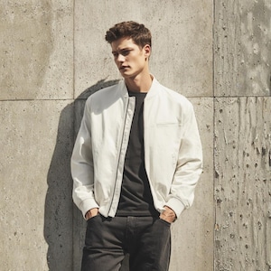 Pode incluir: Casaco bomber branco com fecho de correr, usado sobre uma camiseta preta e jeans cinza escuro. O casaco tem um design simples e limpo. A pessoa está em frente a uma parede de concreto texturizado.