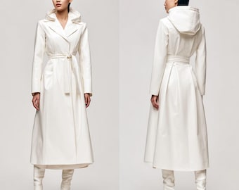 May include: A long, white trench coat with a hood and a matching belt. The coat is knee-length and has a classic design. The model is wearing white boots. The coat is made of a smooth, possibly leather-like material.