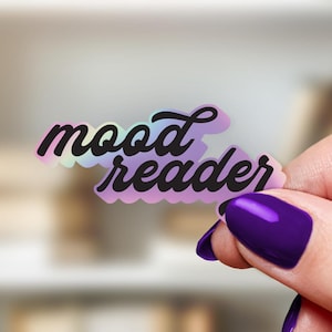 May include: A holographic sticker with the words "mood reader" in a script font. The sticker has a scalloped edge and is held by a hand with purple nail polish. The background is a blurred bookshelf.