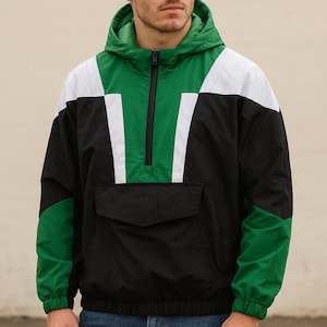 May include: A hooded anorak jacket in black, green, and white. The jacket features a half-zip closure, a front pocket, and color-blocked panels on the shoulders and sleeves. The jacket is worn with blue jeans.