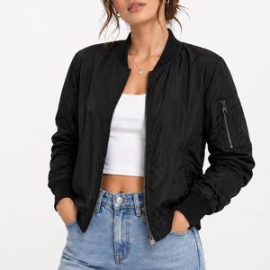 May include: A black bomber jacket with a zipper closure and a zippered pocket on the left sleeve. The jacket has ribbed cuffs and a ribbed hem. The model is wearing a white crop top and blue jeans.