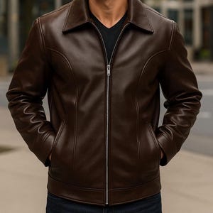 May include: A brown leather jacket with a collared neckline and a silver zipper closure. The jacket has two front pockets and a tailored fit. The model is wearing a black shirt underneath. The jacket is a classic style.