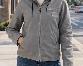 May include: A gray hooded jacket with a zipper closure, two front pockets, and a small logo patch on the chest. The jacket has a drawstring hood and a soft, textured lining. The person is wearing blue jeans.