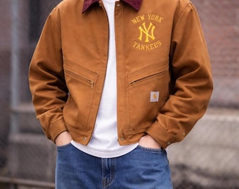 May include: A brown jacket with a burgundy collar and the words "NEW YORK YANKEES" embroidered in yellow. The jacket has two front pockets and a small square logo on the lower right side. The person is wearing blue jeans and a white t-shirt.