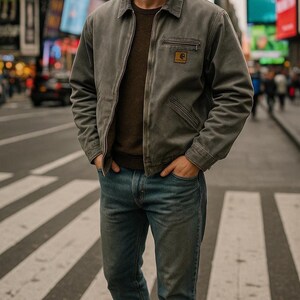 May include: A grey work jacket with a brown sweater and blue jeans. The jacket features a zip closure, a small chest pocket, and a logo patch. The person is standing on a pedestrian crossing.
