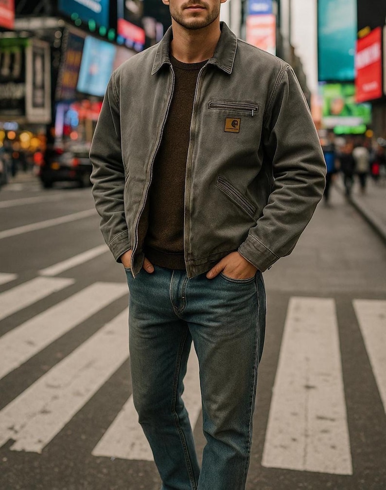 May include: A grey work jacket with a brown sweater and blue jeans. The jacket features a zip closure, a small chest pocket, and a logo patch. The person is standing on a pedestrian crossing.