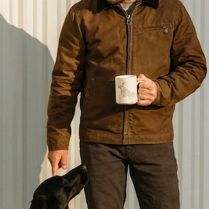 May include: A brown jacket with a black collar and a zipper closure. The person is holding a white mug with a drawing on it. A black dog is looking up at the person.
