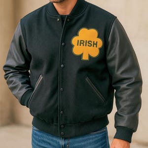 May include: Black varsity jacket with leather sleeves. The jacket features a yellow shamrock patch with the word "IRISH" in black. The jacket has a button-up front, pockets, and a ribbed hem and cuffs. The person is wearing blue jeans.