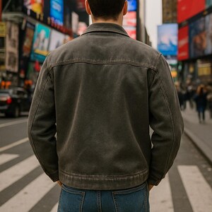 May include: A dark grey denim jacket with a classic collar and button closure. The jacket is paired with blue jeans. The person is standing on a pedestrian crossing in a city setting with bright billboards in the background.