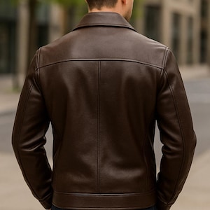 May include: A brown leather jacket with a classic design. The jacket features a collared neckline, a front closure, and a tailored fit. The back of the jacket is visible, showcasing the stitching details. The person is wearing dark blue jeans.
