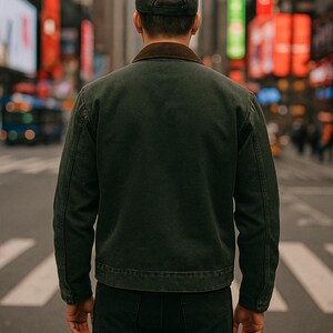 May include: A person wearing a dark green jacket with a brown collar and a dark green baseball cap, standing on a city street. The person is facing away from the camera. The background features city buildings and street lights.