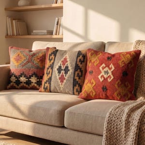 Puede incluir: Tres cojines decorativos con patrones geométricos en tonos rojos, naranjas, negros y grises descansan sobre un sofá beige. Los cojines tienen una apariencia texturizada, agregando interés visual al ambiente de la sala de estar.