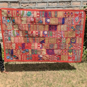 Puede incluir: Un textil rectangular de patchwork con un borde rojo. El textil está compuesto por numerosos parches rectangulares en tonos de rojo, dorado, azul y verde, con intrincados patrones bordados. El textil se sostiene contra una pared de ladrillos y hierba.