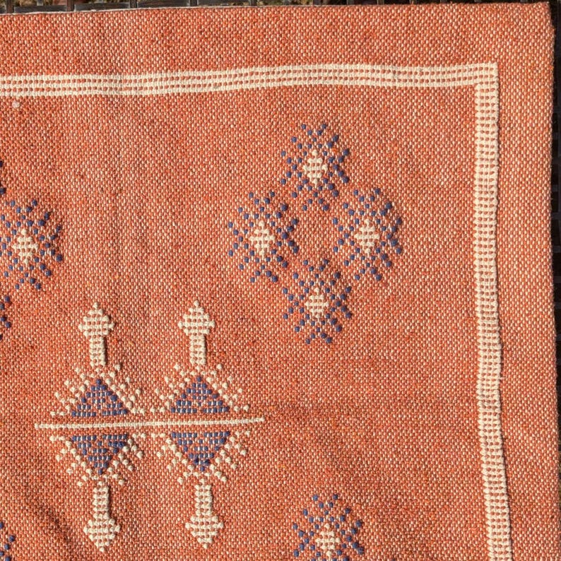 May include: Close-up of a terracotta-colored woven rug with geometric patterns. The rug features a repeating design of diamond and arrow-like shapes in white and blue. A white border frames the edges.