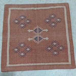 May include: Square terracotta-colored woven pillow cover with a white border. The center features a symmetrical design of diamond and arrow shapes in navy blue and white, creating a geometric pattern. The pillow cover has a textured appearance.