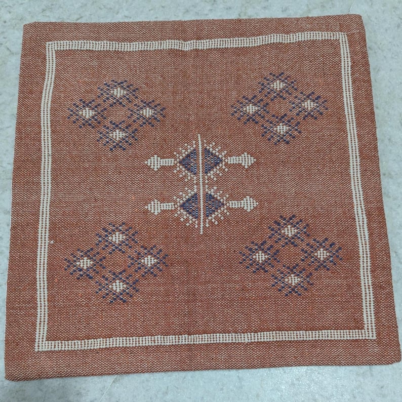 May include: Square terracotta-colored woven pillow cover with a white border. The center features a symmetrical design of diamond and arrow shapes in navy blue and white, creating a geometric pattern. The pillow cover has a textured appearance.
