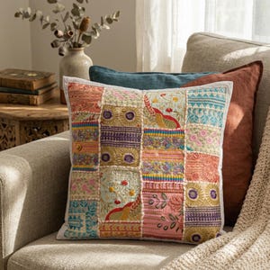 Puede incluir: Un cojín cuadrado decorativo con un diseño de patchwork. El cojín presenta una variedad de cuadrados bordados coloridos en tonos de rosa, azul, naranja y morado. Se coloca en un sofá gris con otros cojines.