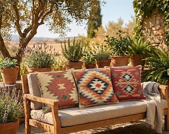 May include: A wooden outdoor bench with three square throw pillows featuring geometric patterns in shades of red, orange, blue, and cream. The bench has a light beige seat cushion and a draped light-colored throw blanket on the right side.