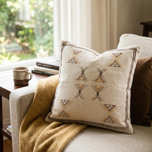 May include: A square, cream-colored decorative pillow with a geometric pattern in brown and gold. The pillow is on a light-colored sofa with a mustard-yellow throw blanket. A brown mug sits on a side table.