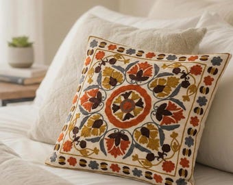 Puede incluir: Un cojín cuadrado con una base de tela color blanco roto y bordados intrincados en tonos naranja quemado, amarillo mostaza, marrón oscuro y azul pizarra. El diseño incluye un motivo floral circular central y un borde decorativo.