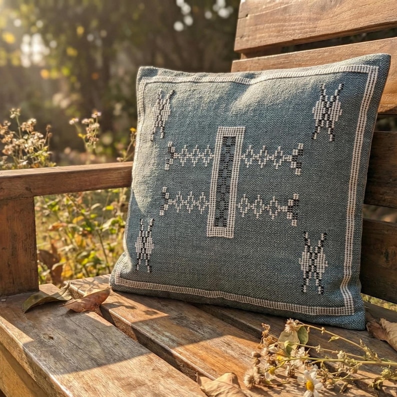 May include: A square, teal-colored decorative pillow with a geometric pattern in white and black. The pillow is placed on a wooden bench outdoors, with sunlight illuminating the scene. The pillow has a textured appearance.