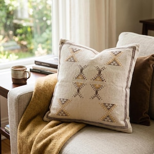 Puede incluir: Cojín decorativo color crema con patrones geométricos en marrón y dorado. El cojín está sobre un sofá de color claro, con una manta amarilla mostaza. Una taza marrón está sobre una mesa auxiliar.