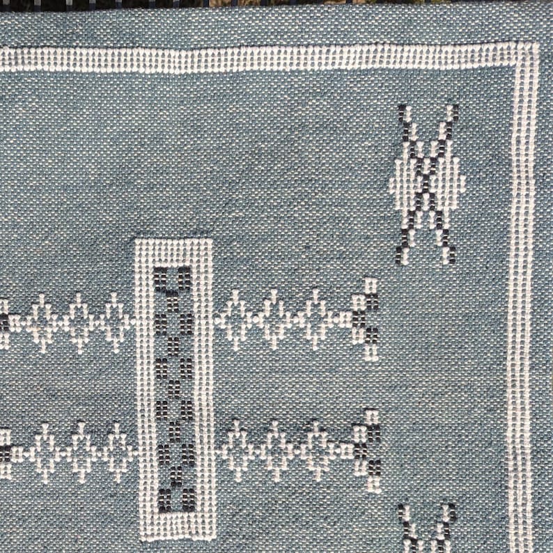 May include: Close-up of a woven textile with a teal background. The fabric features white and black geometric patterns, including diamond and rectangular shapes. White borders and accents frame the design, creating a textured, decorative piece.