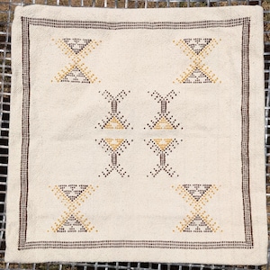 May include: Cream-colored square textile with a brown border and geometric patterns. The design features repeating shapes in brown and yellow, creating a symmetrical arrangement. The textile is likely a decorative item, such as a pillow cover or small rug.