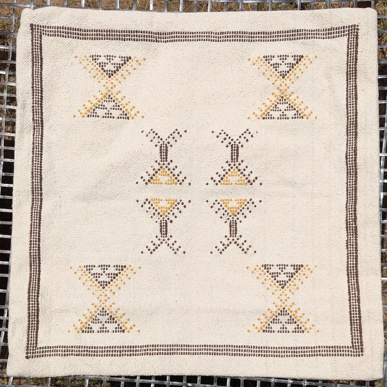 May include: Cream-colored square textile with a brown border and geometric patterns. The design features repeating shapes in brown and yellow, creating a symmetrical arrangement. The textile is likely a decorative item, such as a pillow cover or small rug.