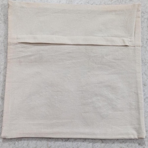 May include: A square, off-white pillow cover made of a textured fabric. The back of the pillow cover has a flap opening. The pillow cover is laying on a light gray surface.