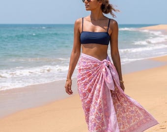 May include: A woman wearing a navy blue bikini top and a pink and white floral sarong tied at the waist. The sarong features a repeating floral pattern and striped borders. The background shows a sandy beach and the ocean.