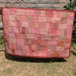 May include: A patchwork quilt in shades of pink, red, and peach. The quilt is made of numerous fabric squares, each with a unique pattern or texture. The edges are trimmed with a darker red border. The quilt is held up against a brick wall and green grass.