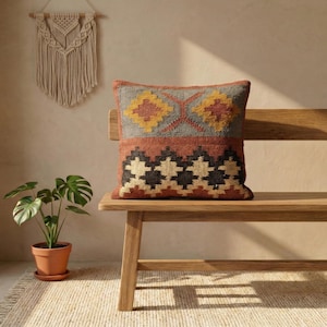 Op de afbeelding: Een vierkant sierkussen met een geweven geometrisch patroon in tinten mosterdgeel, terracotta oranje, antracietgrijs en crème. Het ontwerp bevat een bovengedeelte met ruitmotieven en een ondergedeelte met een herhalend zigzagpatroon.