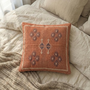 May include: Square terracotta-colored decorative pillow with a geometric pattern in dark blue and cream. The pillow is placed on a bed with neutral-colored bedding and a knitted throw blanket. The pillow has a cream-colored border.