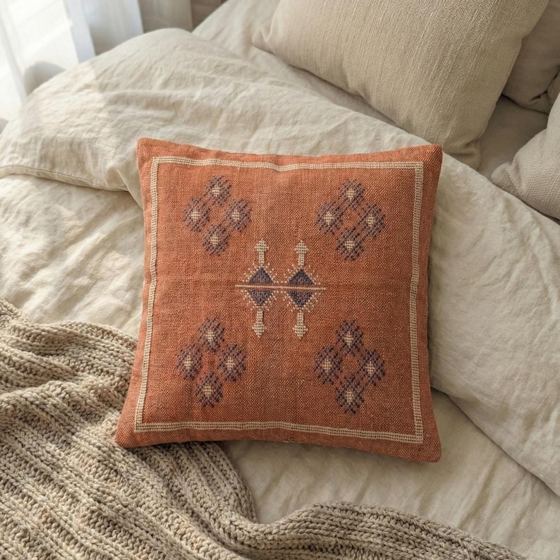May include: Square terracotta-colored decorative pillow with a geometric pattern in dark blue and cream. The pillow is placed on a bed with neutral-colored bedding and a knitted throw blanket. The pillow has a cream-colored border.