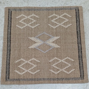 May include: A square, woven textile in shades of tan, with a geometric pattern in white and light blue. The design features four diamond-shaped motifs and a central, stylized diamond. The border is a black and white striped pattern.