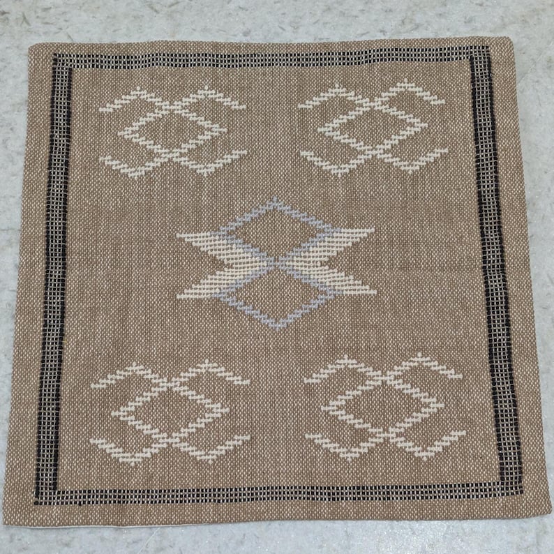 May include: A square, woven textile in shades of tan, with a geometric pattern in white and light blue. The design features four diamond-shaped motifs and a central, stylized diamond. The border is a black and white striped pattern.