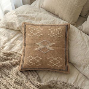 May include: A square, tan-colored decorative pillow with a geometric pattern in white and gray. The pillow has a textured appearance and a dark border. The pillow is placed on a bed with neutral-colored bedding.
