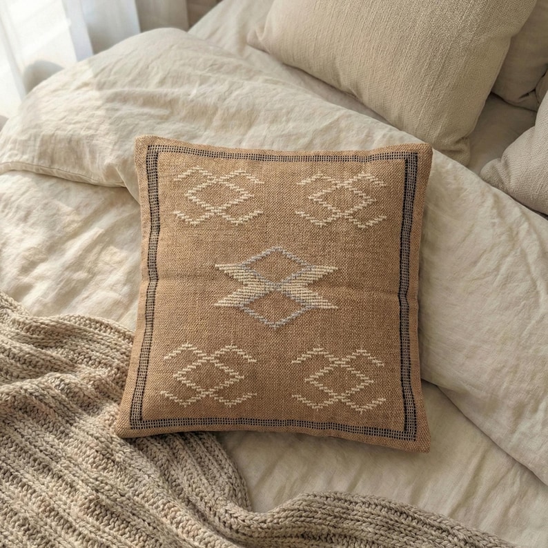 May include: A square, tan-colored decorative pillow with a geometric pattern in white and gray. The pillow has a textured appearance and a dark border. The pillow is placed on a bed with neutral-colored bedding.