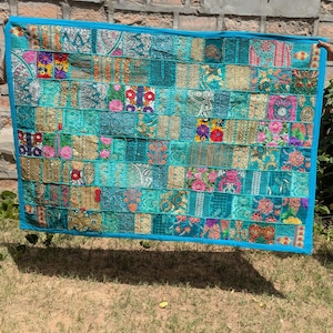 Puede incluir: Un edredón rectangular de patchwork con una vibrante mezcla de telas turquesa, doradas y rosas. El edredón presenta un borde azul y está compuesto por numerosos parches rectangulares con diseños florales y geométricos. El edredón se sostiene contra una pared de ladrillos y hierba.
